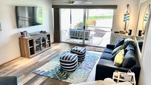 Ein modernes Wohnzimmer mit Flachbildfernseher, Holzboden, einer Sofagarnitur, gestreiften Hocker und einem großen Fenster mit Blick ins Freie.