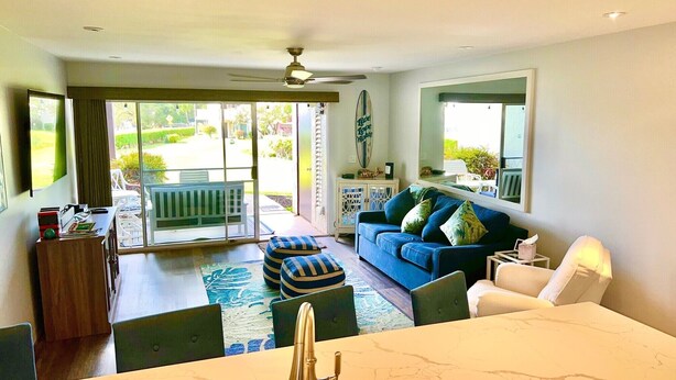 Ein geräumiges Wohnzimmer mit einer großen Glasschiebetür, die auf eine Terrasse führt, ein blaues Sofa mit Kissen, ein weißer Sessel und ein Essbereich mit einem Marmortisch.