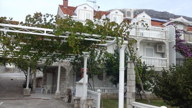 A residential building with a stone facade, white balconies, and a pergola with climbing plants.