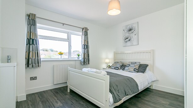 A bedroom with a bed, a window with curtains, a radiator, a wall-mounted light fixture, and a flower vase. - Denby Lodge (Letchworth)