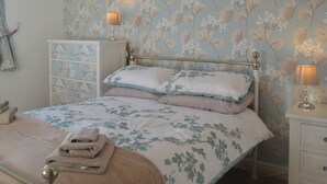 Een slaapkamer met bloemenbehang, een bed met twee kussens, een nachtkastje met een lamp, een dressoir en een stapel handdoeken.