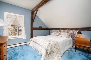 Une chambre avec un plafond en pente, un lit recouvert d’une literie à motifs floraux, un banc en bois et une fenêtre avec des rideaux.