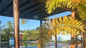 Un espace extérieur aménagé, abrité et doté de mobilier en bois, avec une piscine en arrière-plan.