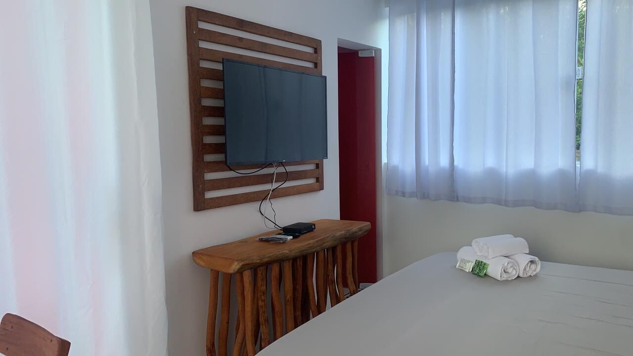 A hotel room with a bed, a TV mounted on a wooden panel, a small wooden table with a phone and a remote, and a window with sheer curtains.