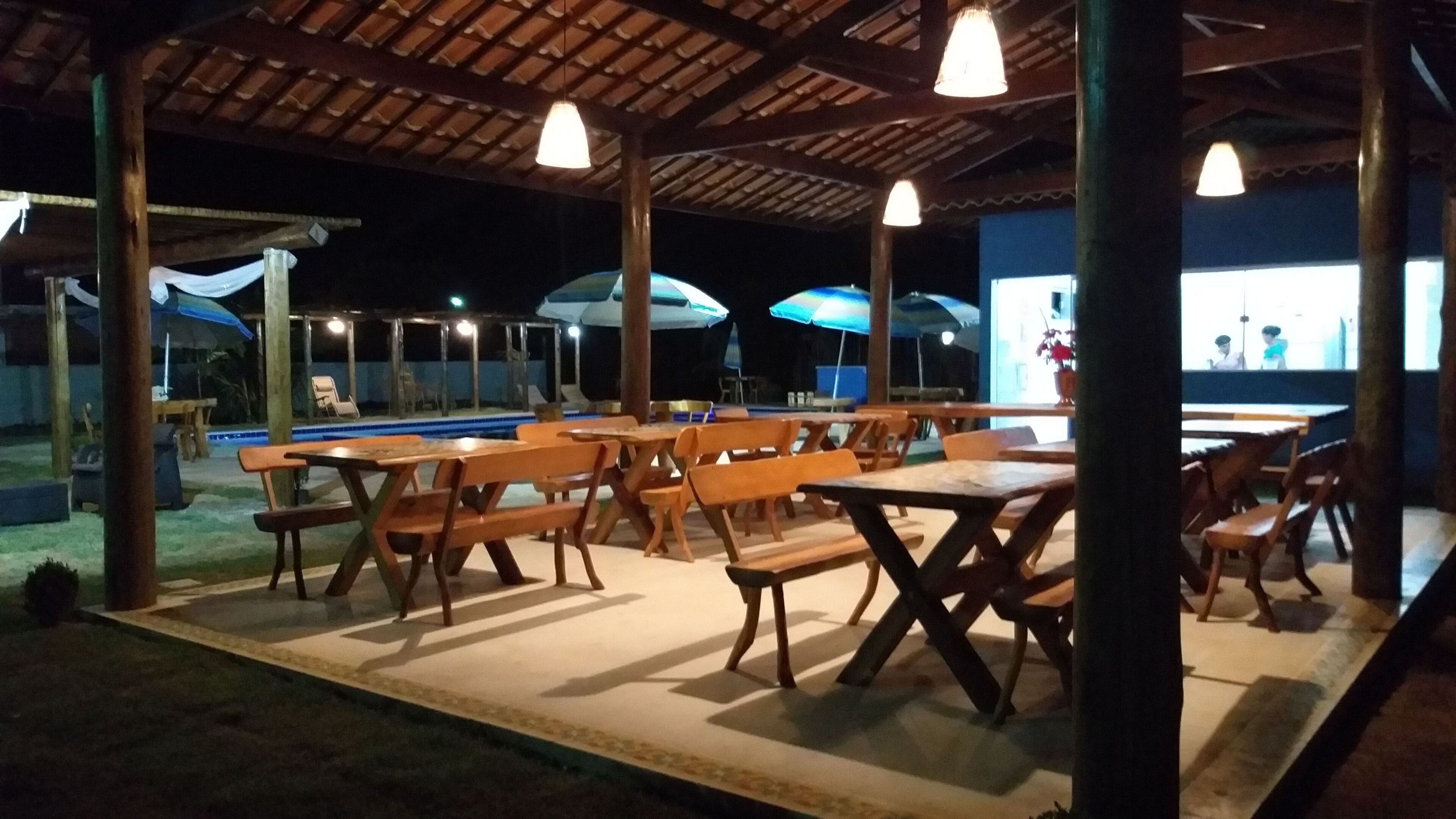An outdoor dining area with wooden tables and benches, umbrellas, and a pool in the background.