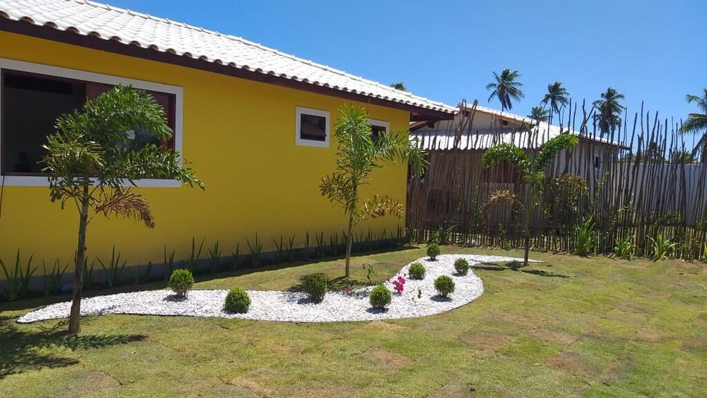Un bâtiment jaune avec un toit en tuiles, entouré d’un jardin où poussent de petits arbres et où l’on trouve des allées recouvertes de gravier blanc.