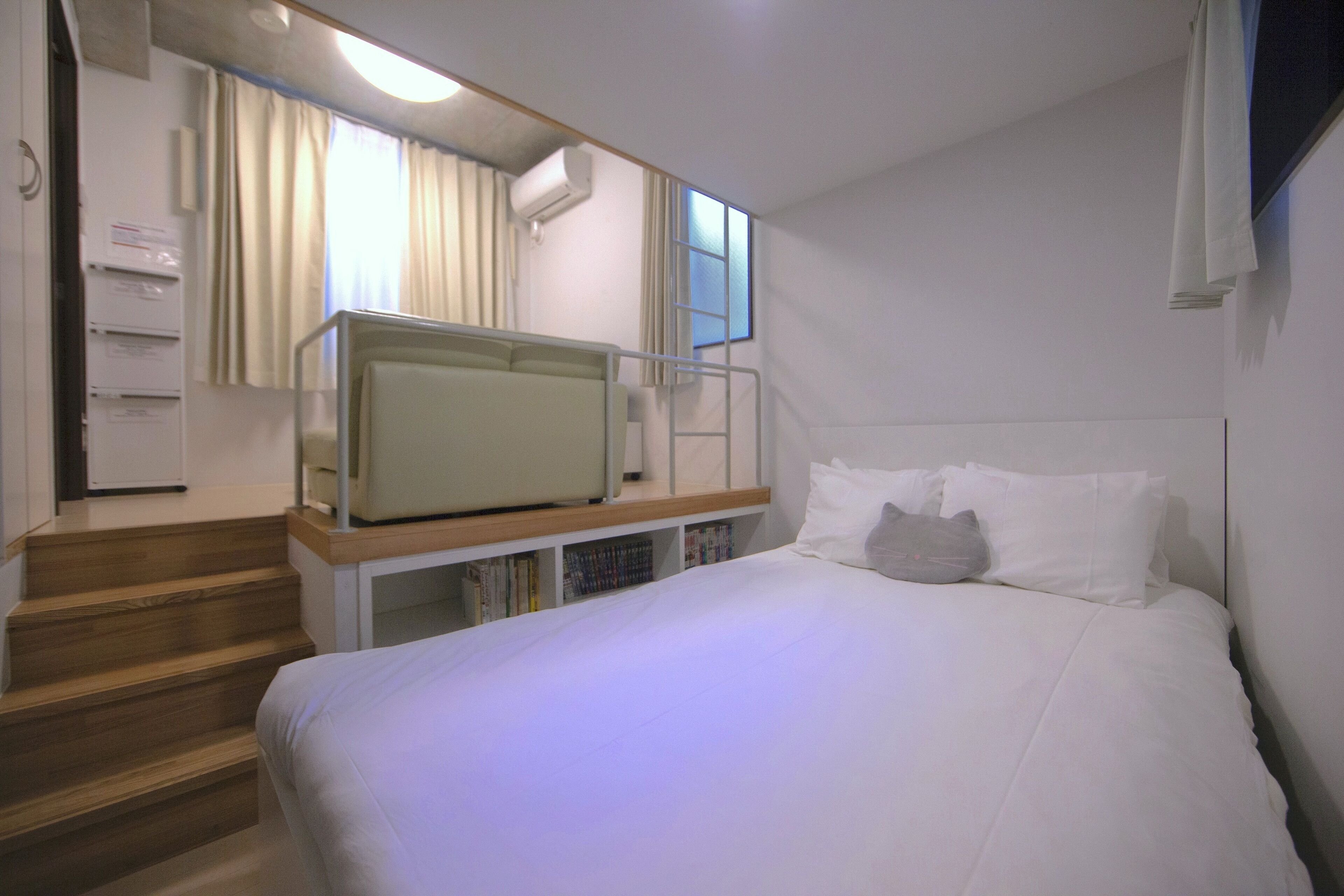 A hotel room with a bed, a desk with a chair, a bookshelf, and a window with curtains.