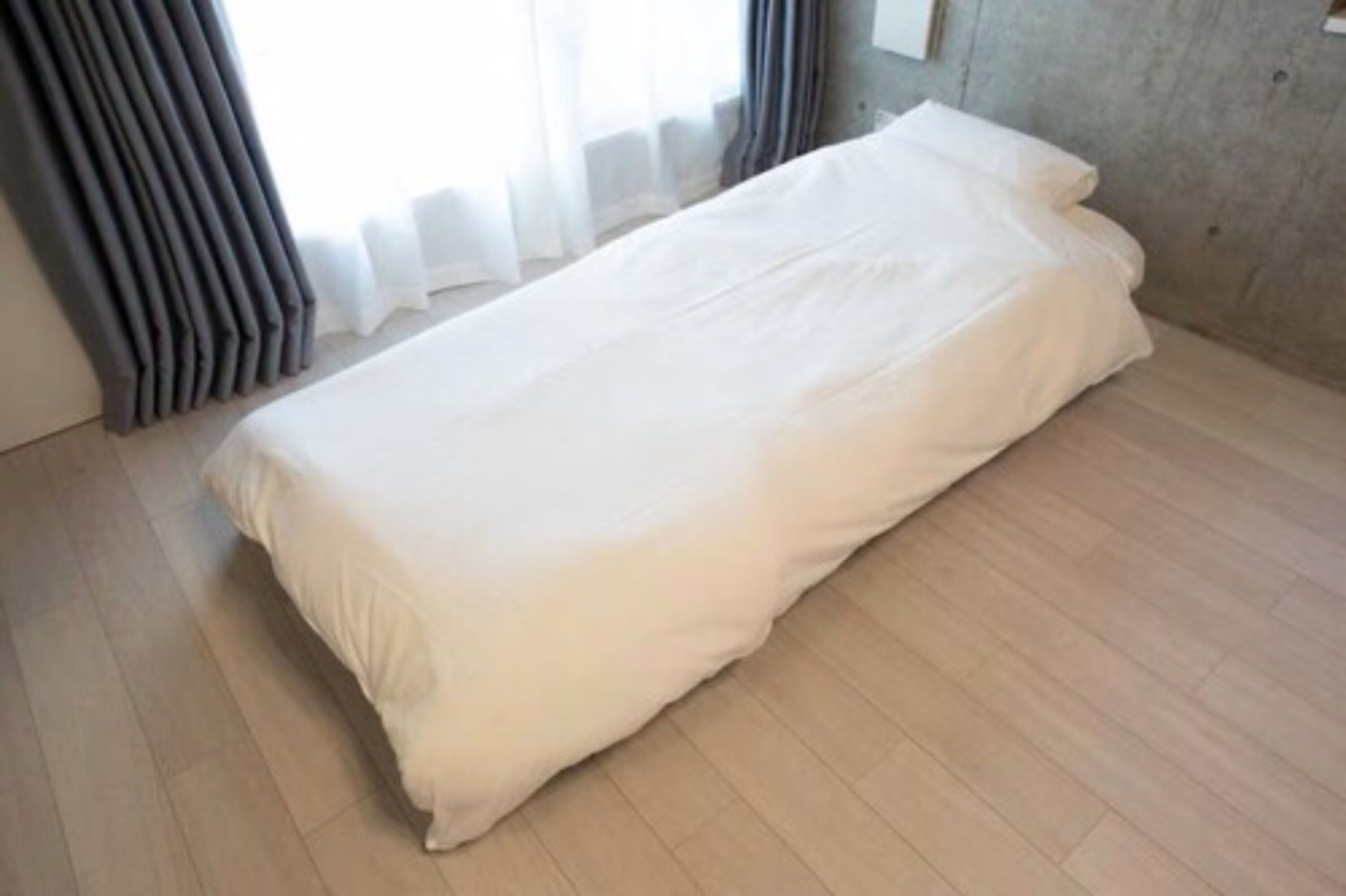 Ein Einzelbett mit weißer Bettwäsche in einem Zimmer mit hellem Holzboden und einer Betonwand.
