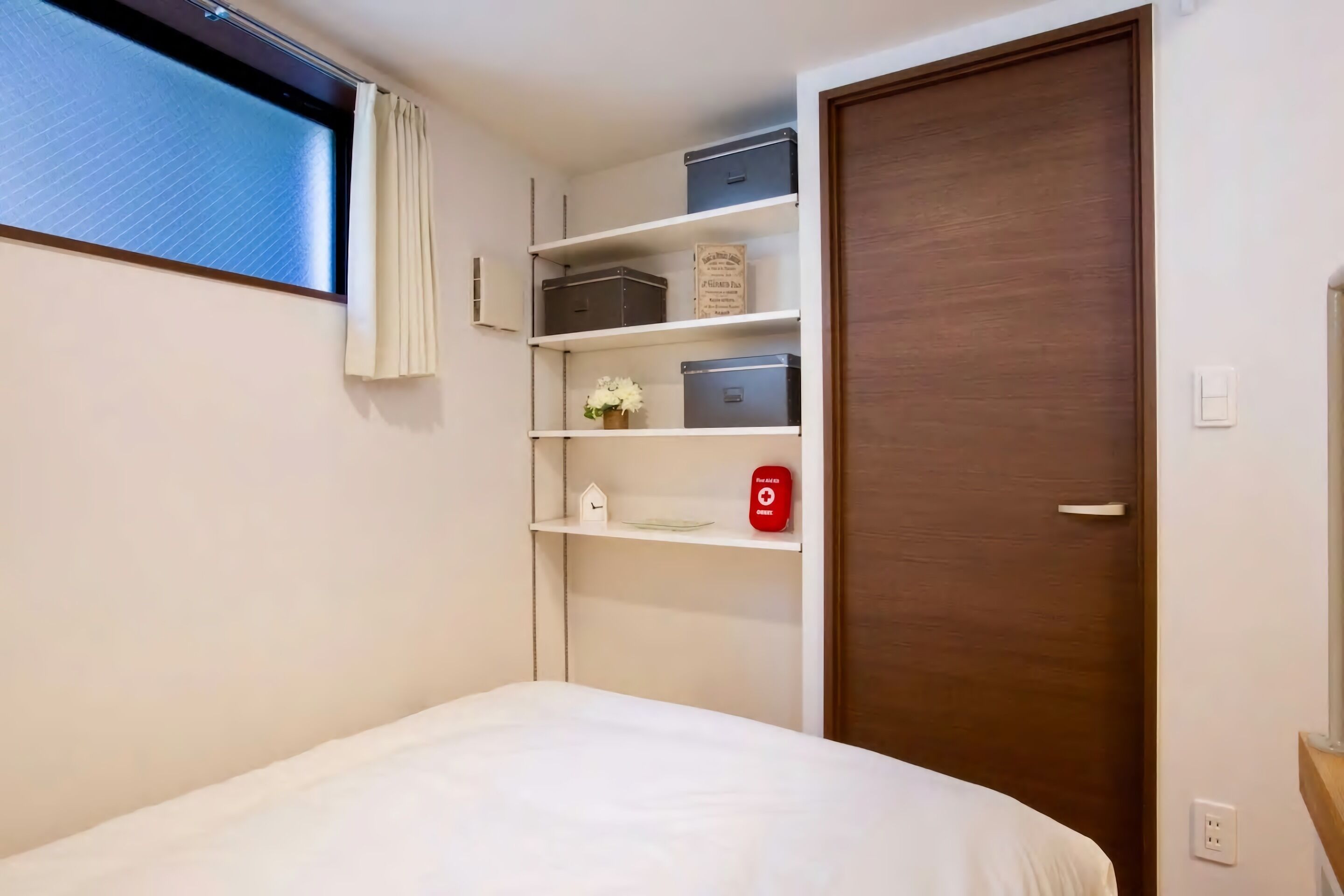A bedroom with a bed, a window with curtains, a wooden door, and built-in shelves with storage boxes and a small clock.