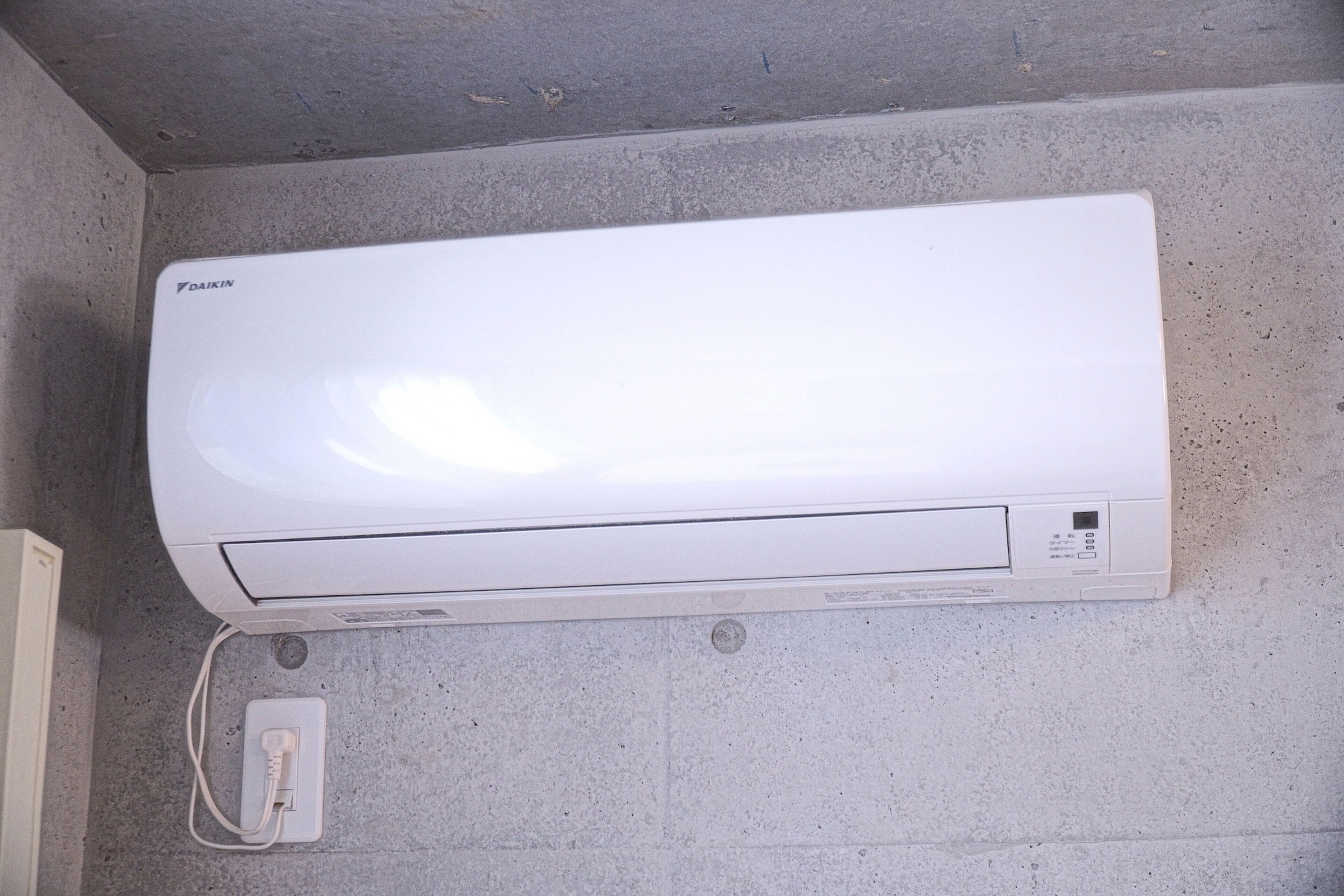 A white Daikin air conditioner mounted on a concrete wall with a power outlet below it.