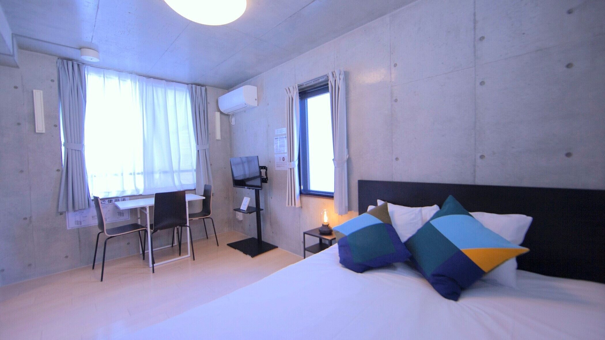A hotel room with a bed, two windows, a desk with a chair, a TV, and a small table with a lamp.