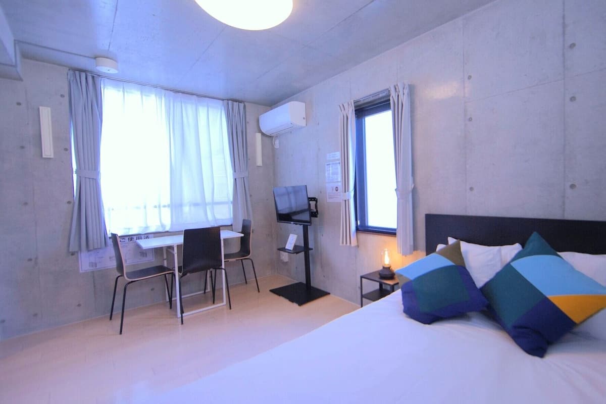 A hotel room with a bed, two windows, a desk with a chair, a TV, and a small table with a lamp.