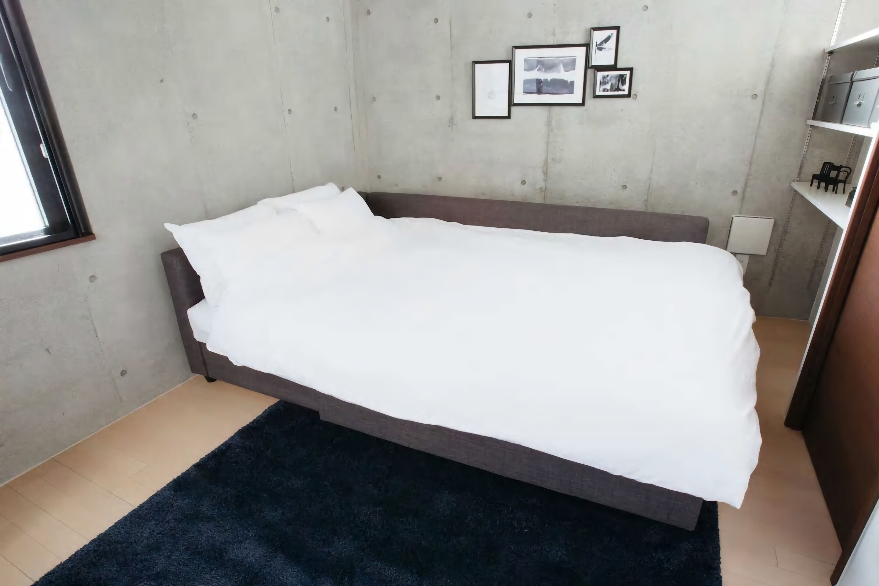 Un dormitorio con una cama, una alfombra oscura, una ventana y una pared con cuadros.