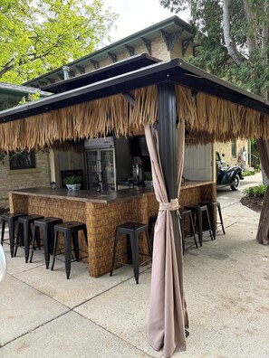 An outdoor bar area with a thatched roof, bar stools, and a vehicle parked nearby.