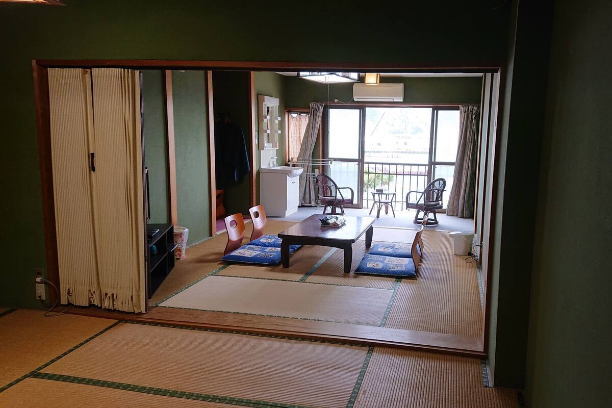 Une pièce de style japonais traditionnel, avec un sol recouvert de tatamis, une petite table basse entourée de coussins et une porte coulissante donnant sur un balcon avec vue.