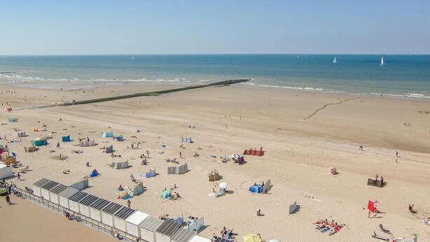 Une plage avec une longue jetée qui s’avance dans l’eau, de nombreux parasols et des gens qui se détendent sur le sable.