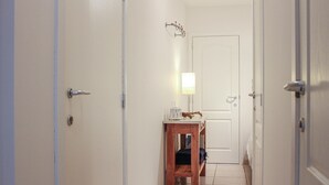 Un couloir étroit aux murs blancs, un banc en bois avec une lampe, et une porte au fond.