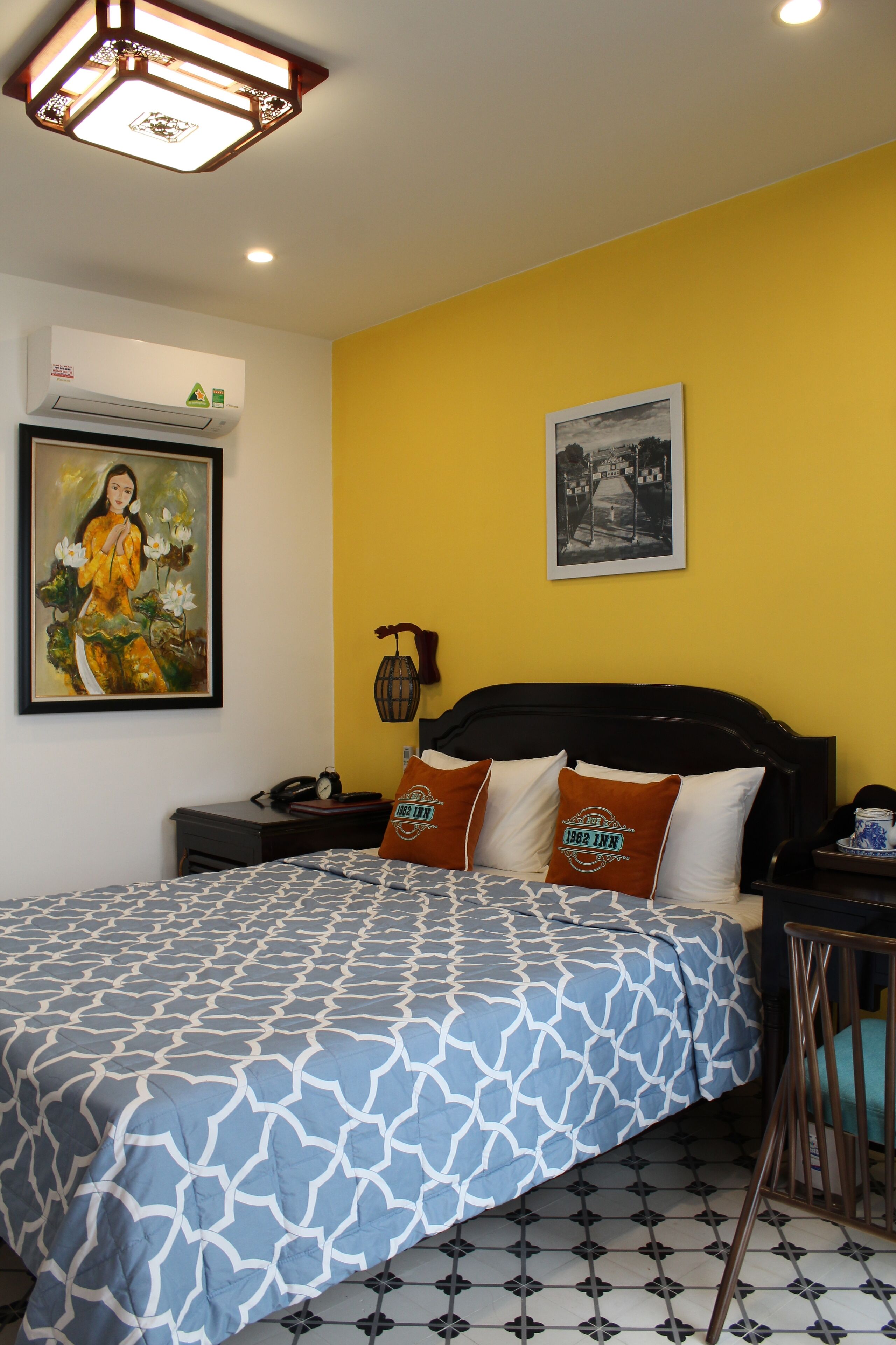 A bedroom with a bed, a nightstand, a wall-mounted air conditioner, a framed picture, and a wall-mounted light fixture.