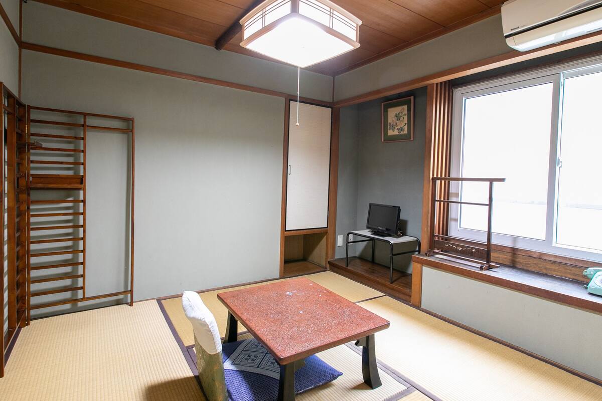A traditional Japanese room with tatami flooring, a low table, a sliding door, and a window with a view.
