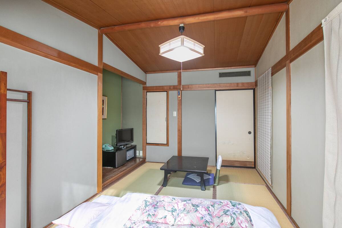 A traditional Japanese-style room with tatami flooring, a bed with a floral bedspread, a television on a low table, and sliding doors.