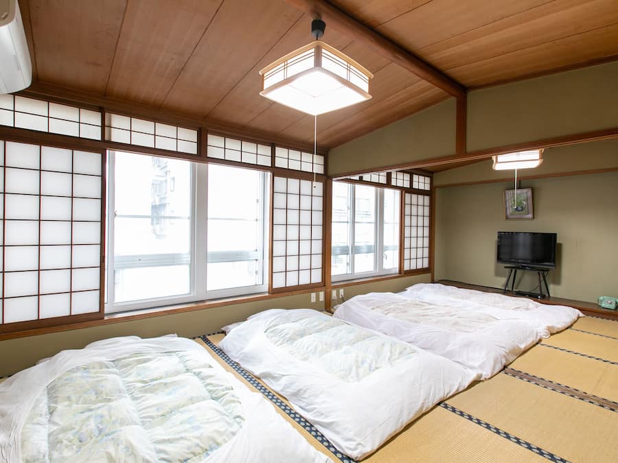 Una habitación tradicional japonesa con suelo de tatami, futón, puertas correderas shoji y un televisor sobre un mueble.