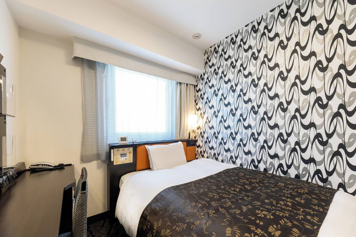A hotel room with a bed, a desk with a telephone, a window with curtains, and a patterned wall.