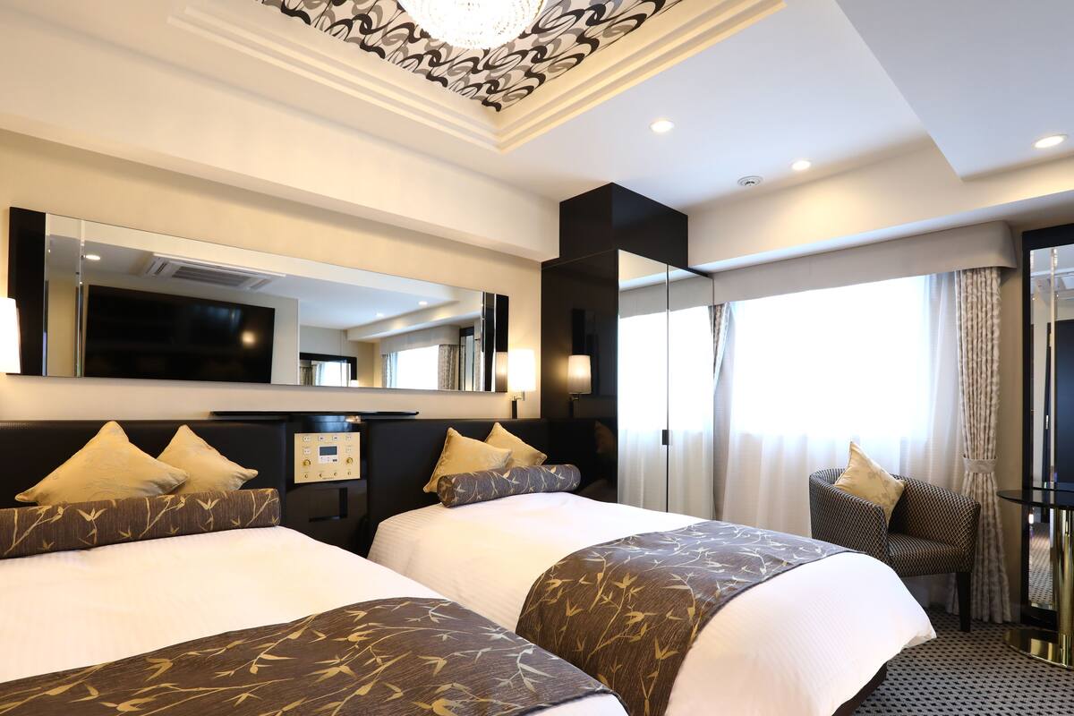 A hotel room with two beds, a large mirror, a TV, and a chair by the window.