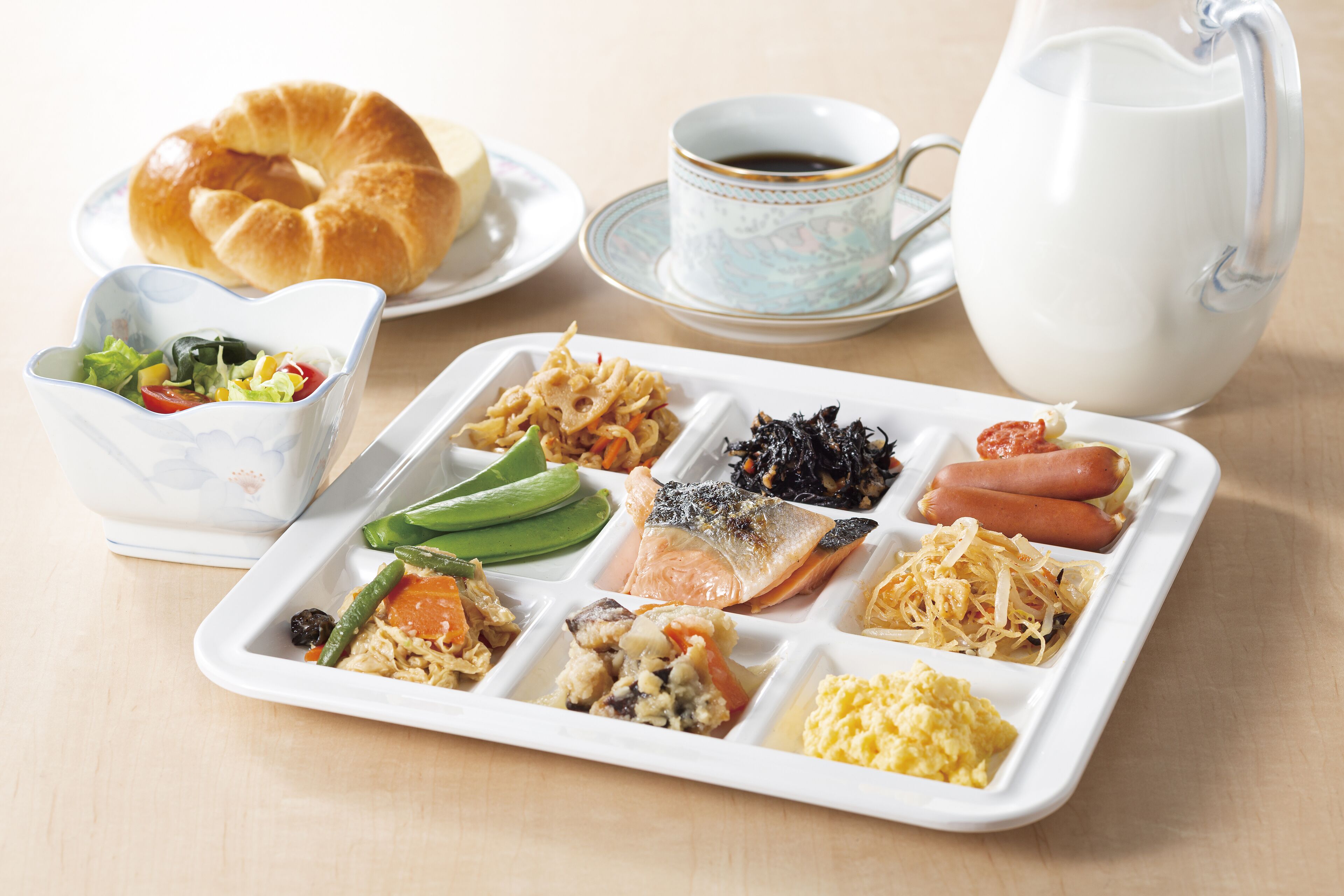 Un plateau repas contenant divers aliments, un croissant, une tasse de café et une carafe de lait.