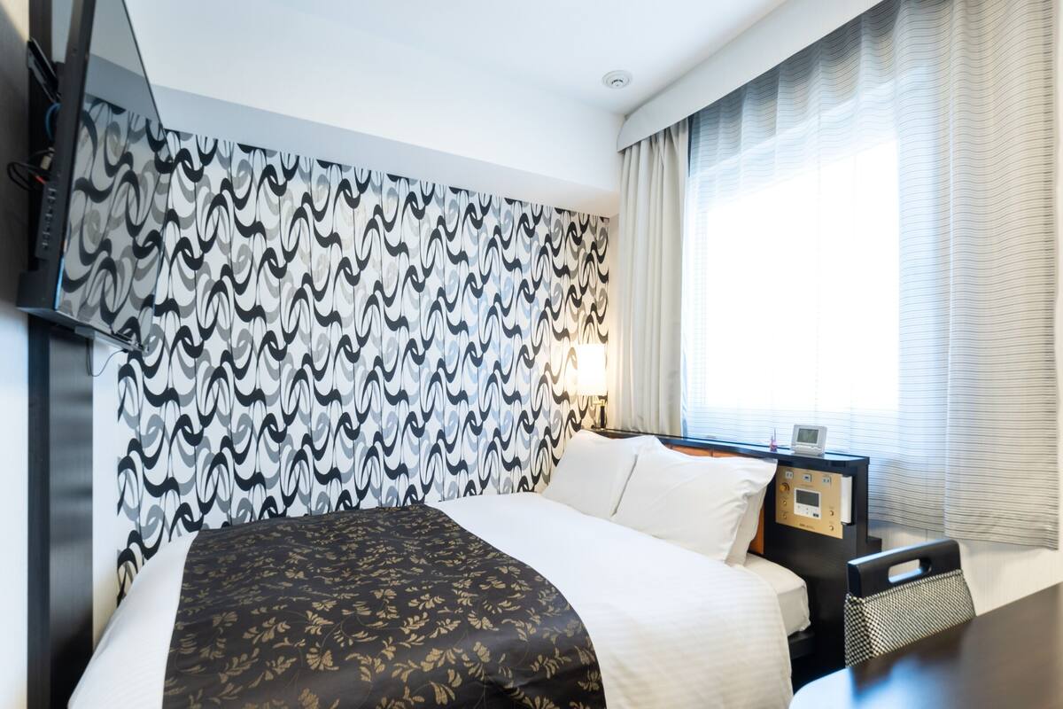A hotel room with a bed, a bedside table with a lamp, a TV mounted on the wall, and a window with curtains.