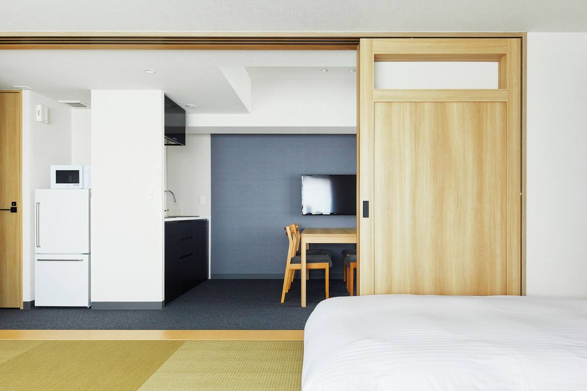 A modern hotel room with a bed, a desk with a chair, a television, and a kitchenette.