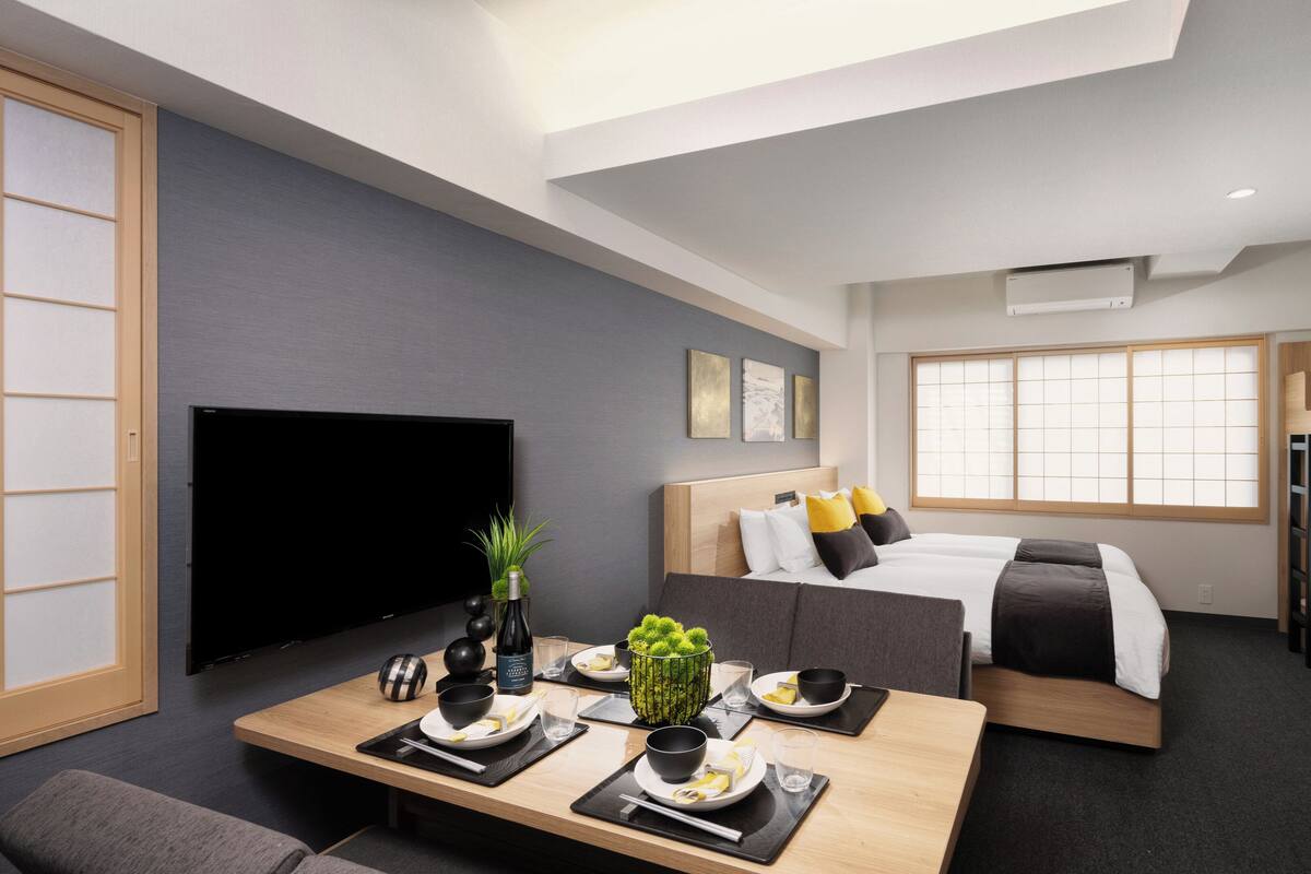 A modern hotel room with a flat-screen TV, a bed with a wooden headboard, a dining area with a table set for two, and a window with a view of the city.
