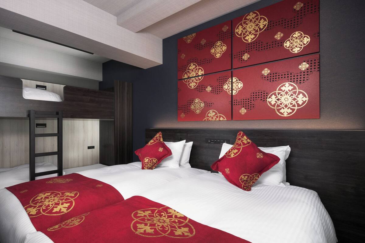 A hotel room with a bunk bed, a large bed with red and gold bedding, and a wall with red panels featuring gold designs.