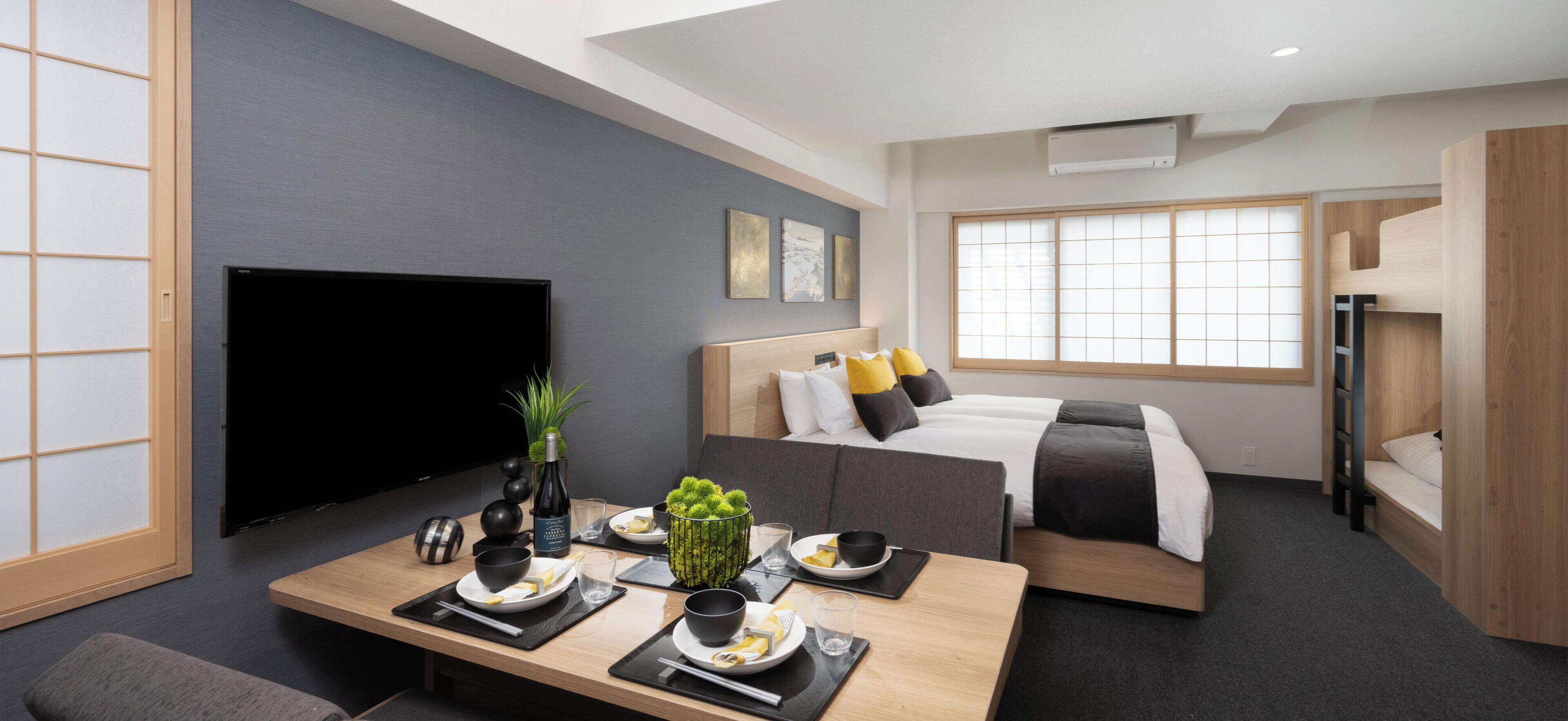 A modern hotel room with a flat-screen TV, a dining table set for two, a bed with a wooden headboard, and a large window with a view.