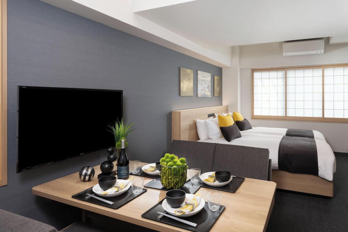 A modern hotel room with a flat-screen TV, a dining table set for two, a bed with a wooden headboard, and a large window with a view.