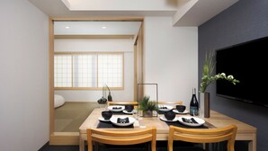 Ein Essbereich mit einem Holztisch, gedeckt für vier Personen, einem an der Wand befestigten Fernseher und Blick auf einen traditionellen japanischen Raum mit Schiebetüren.