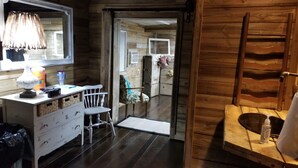 Interior de una cabaña de madera con un aparador blanco, una silla, una escalera de madera y una puerta que da a otra habitación.