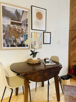 A dining area with a wooden table, chairs, and a vase of white flowers. There are framed pictures on the wall.