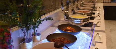 Un bufé con diversas opciones de comida, incluyendo tazones de sopa y otros platos, dispuesto en una mesa con barrera de vidrio.