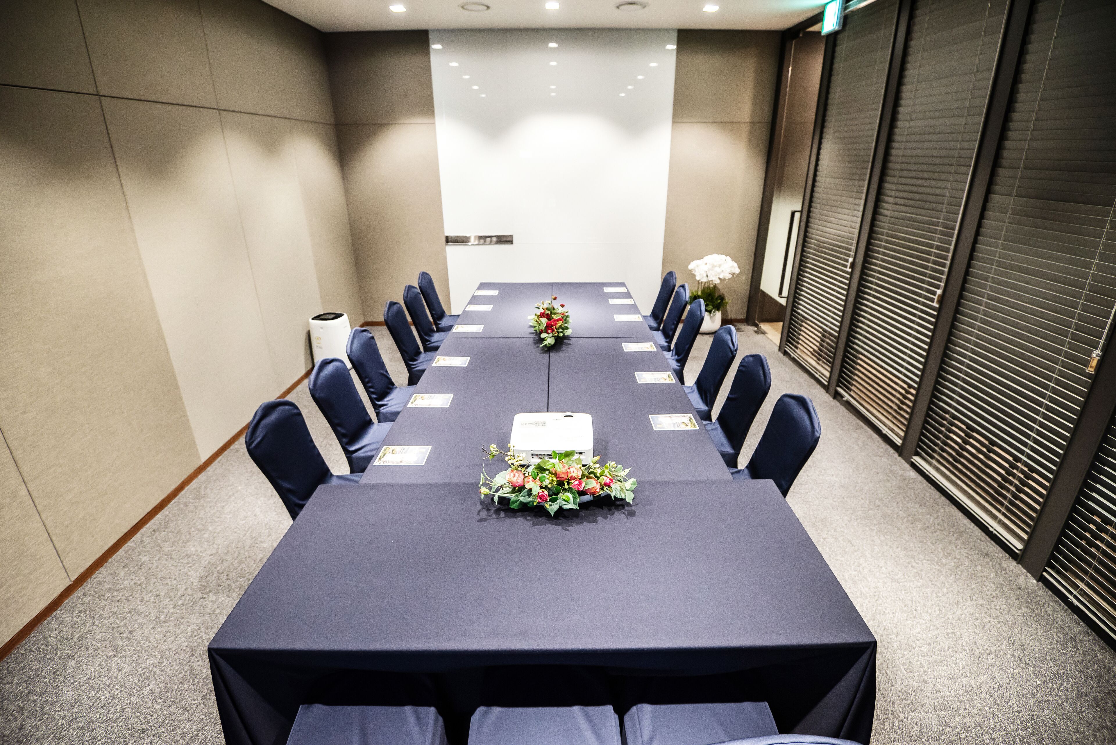 Un salón de conferencias con una larga mesa, sillas, un proyector y una decoración floral.
