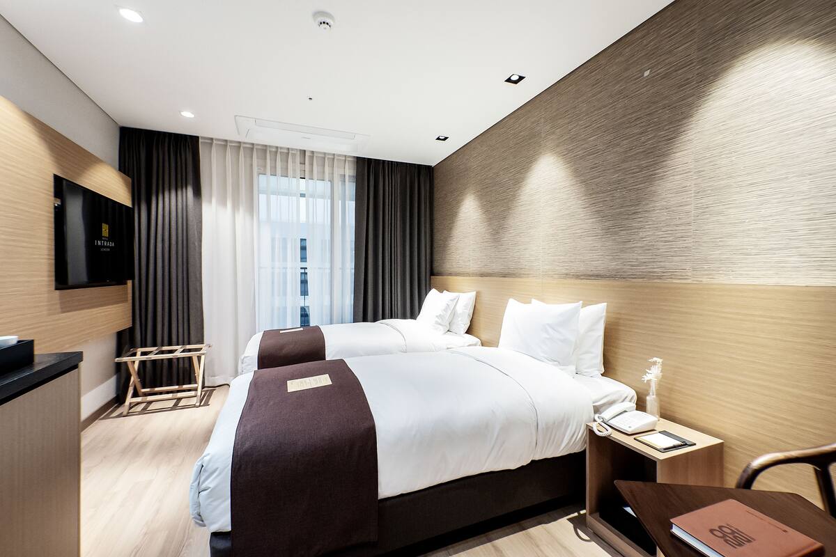 A modern hotel room with two beds, a flat-screen TV, a bedside table with a lamp, and a view of the city through large windows.