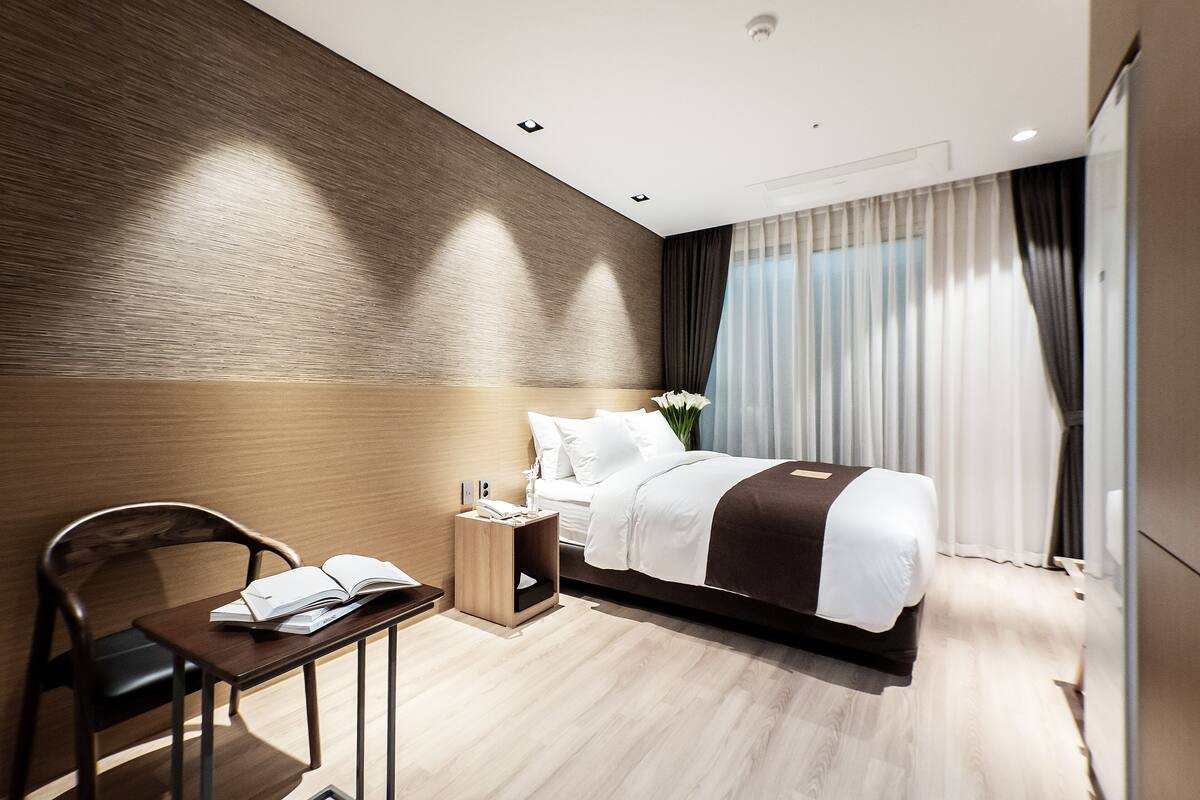 A modern hotel room with a wooden accent wall, a neatly made bed, a small bedside table with a book, and a chair.