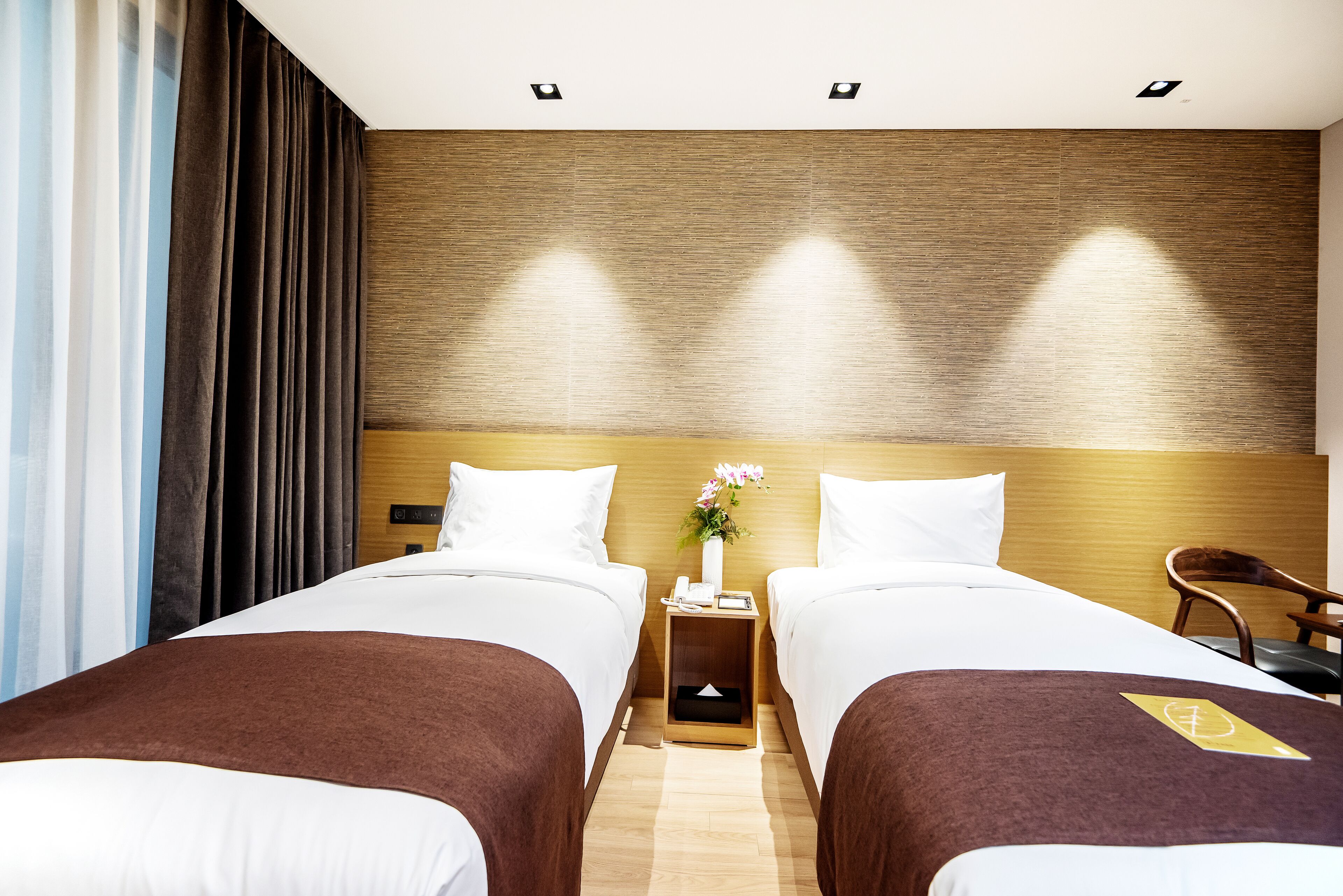 A hotel room with two beds, a wooden headboard, a nightstand with a phone and a vase of flowers, and a chair.