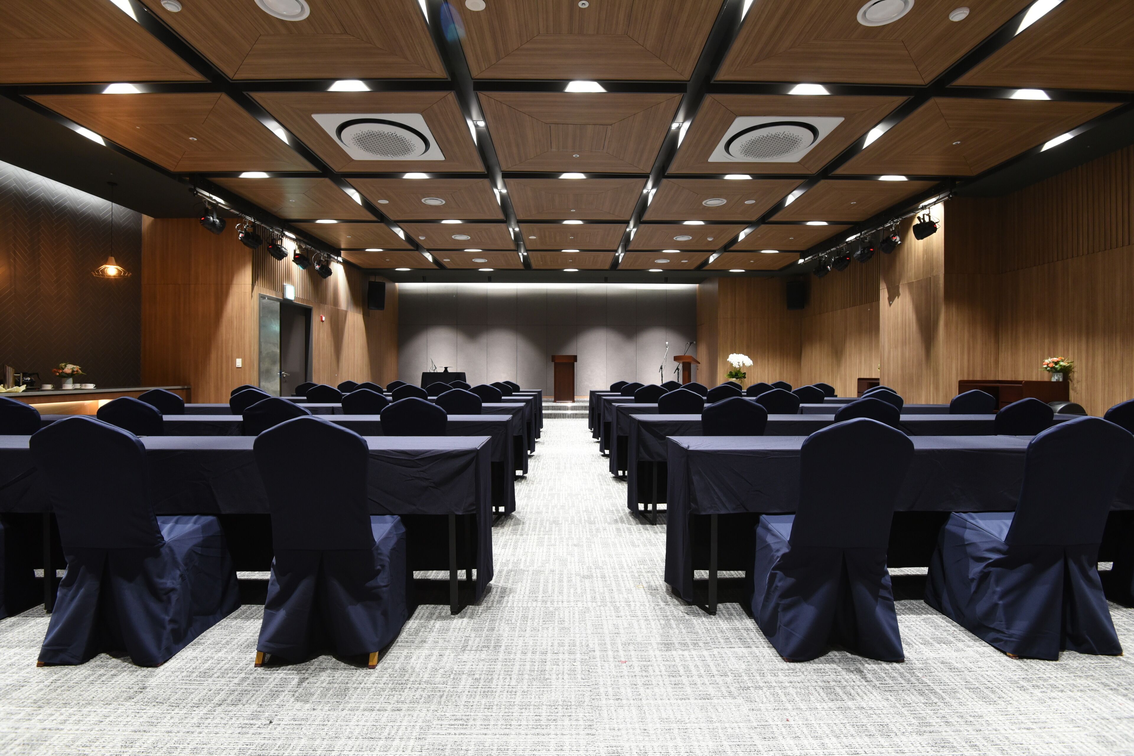 A conference room with rows of tables and chairs, wooden paneling, and ceiling lights.