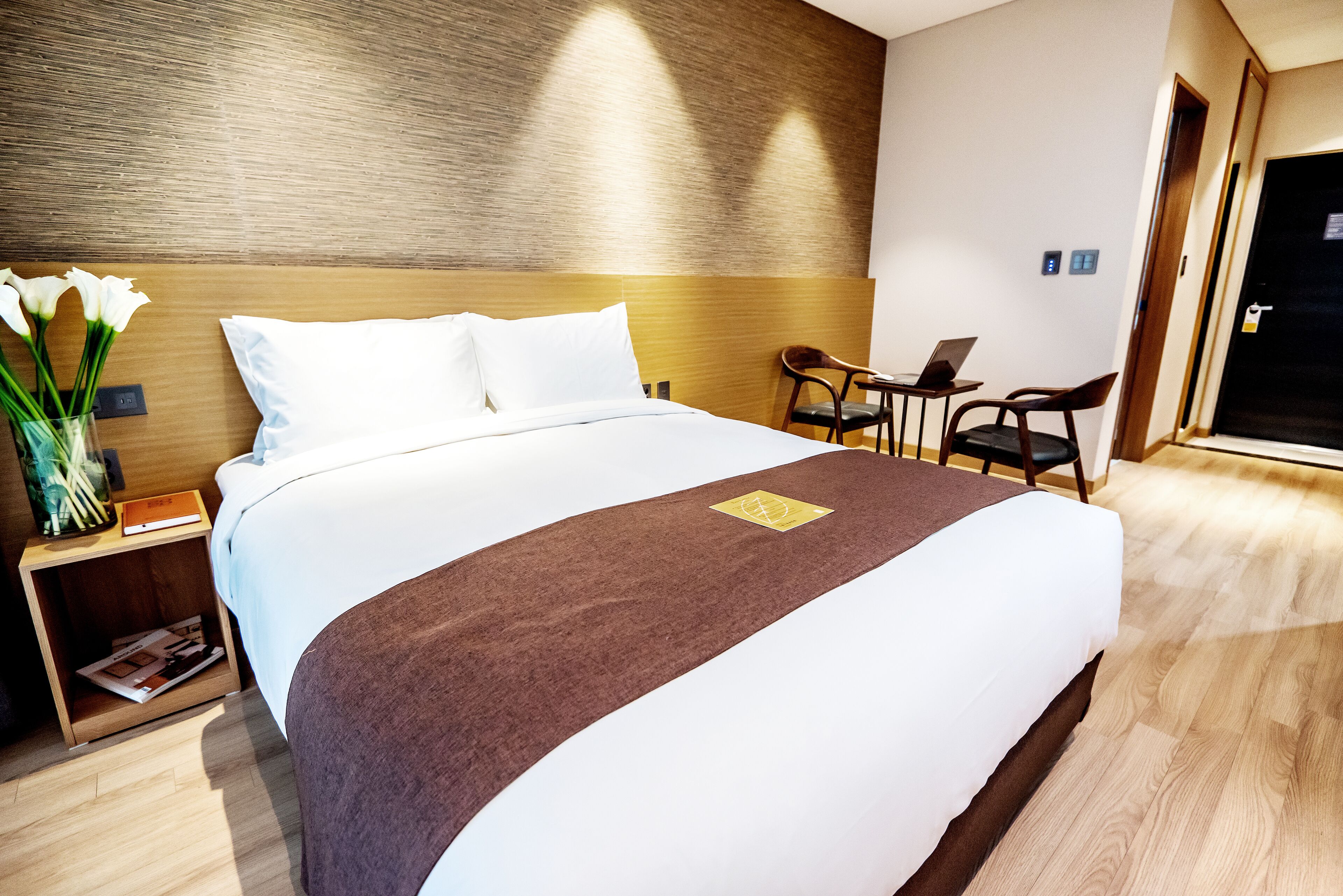 A neatly made bed with a brown runner, a small table with a laptop, and a chair in a modern hotel room.
