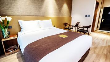 A neatly made bed with a brown runner, a small table with a laptop, and a chair in a modern hotel room.
