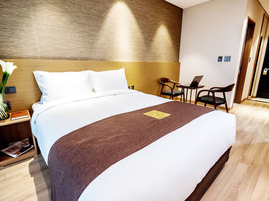 A neatly made bed with a brown runner, a small table with a laptop, and a chair in a modern hotel room.