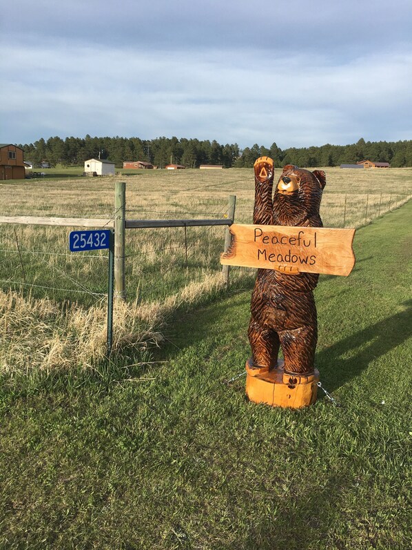 Una escultura de madera de un oso con un letrero que dice "Peaceful Meadows" en un campo cubierto de hierba, con una cerca y un letrero azul que muestra el número 25432.