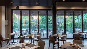 A modern dining area with tables and chairs, large windows, and a view of greenery outside. - Xotel (Khon Kaen)