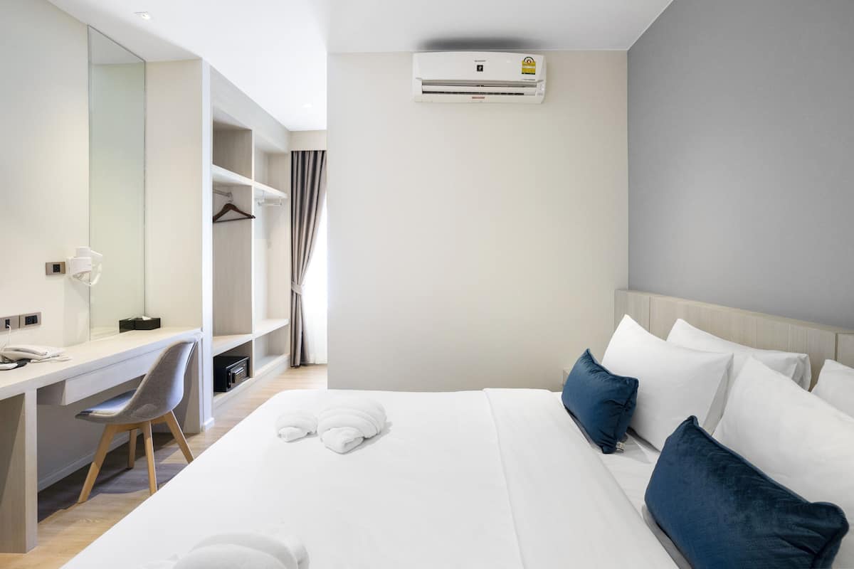 A hotel room with a bed, a desk, a chair, a wardrobe, and an air conditioning unit.