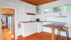 Une cuisine compacte avec des meubles blancs, un plan de travail en bois et un petit réfrigérateur. Une fenêtre offre une vue sur la verdure à l’extérieur, et un extincteur est fixé au mur.